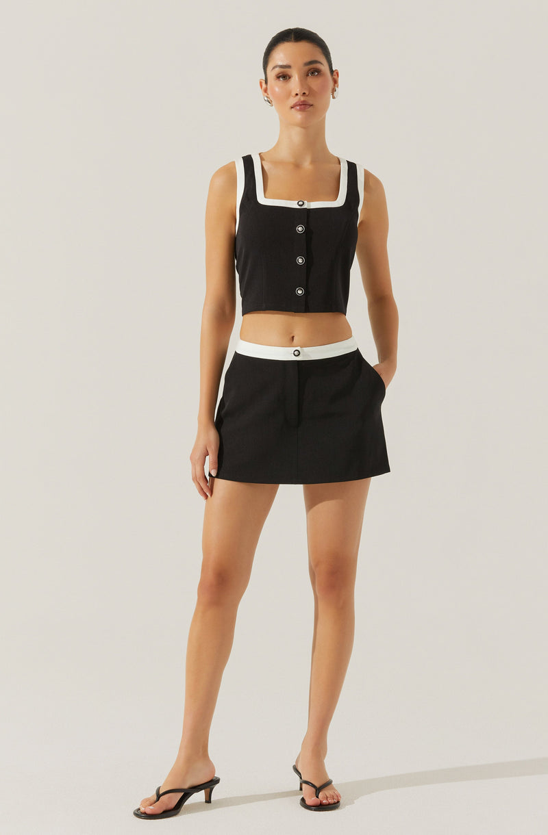 A woman is wearing a black sleeveless crop top with white trim along the square neckline and armholes, featuring black buttons down the front. She pairs the top with a matching black mini skirt that has a white waistband and white button detail at the center. She completes her outfit with black open-toe high-heeled sandals.