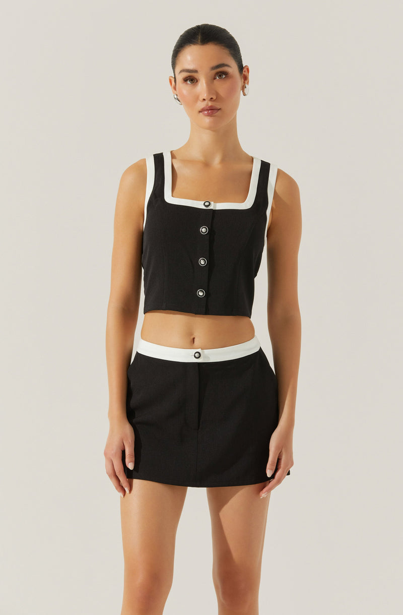 A woman is wearing a black sleeveless cropped top with white trim around the neckline, sleeve edges, and button placket, paired with a matching black mini skirt that has a white waistband with a single black button.