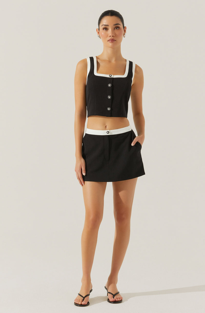 A young woman is wearing a sleeveless black crop top with white trimmed accents around the square neckline and armholes. The top features a row of buttons down the front. She pairs it with a black mini skirt that has a white waistband and a front button detail. She completes her outfit with black strappy sandals.