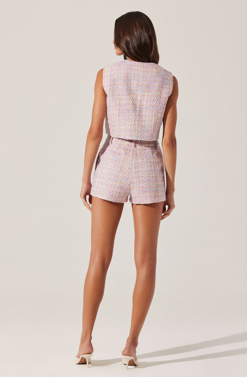 A woman is wearing a sleeveless, pastel-colored, woven textured two-piece outfit consisting of a crop top and high-waisted shorts. The fabric has a multicolored, tweed-like appearance with shades of pink, purple, and cream. She is also wearing white high-heeled shoes.