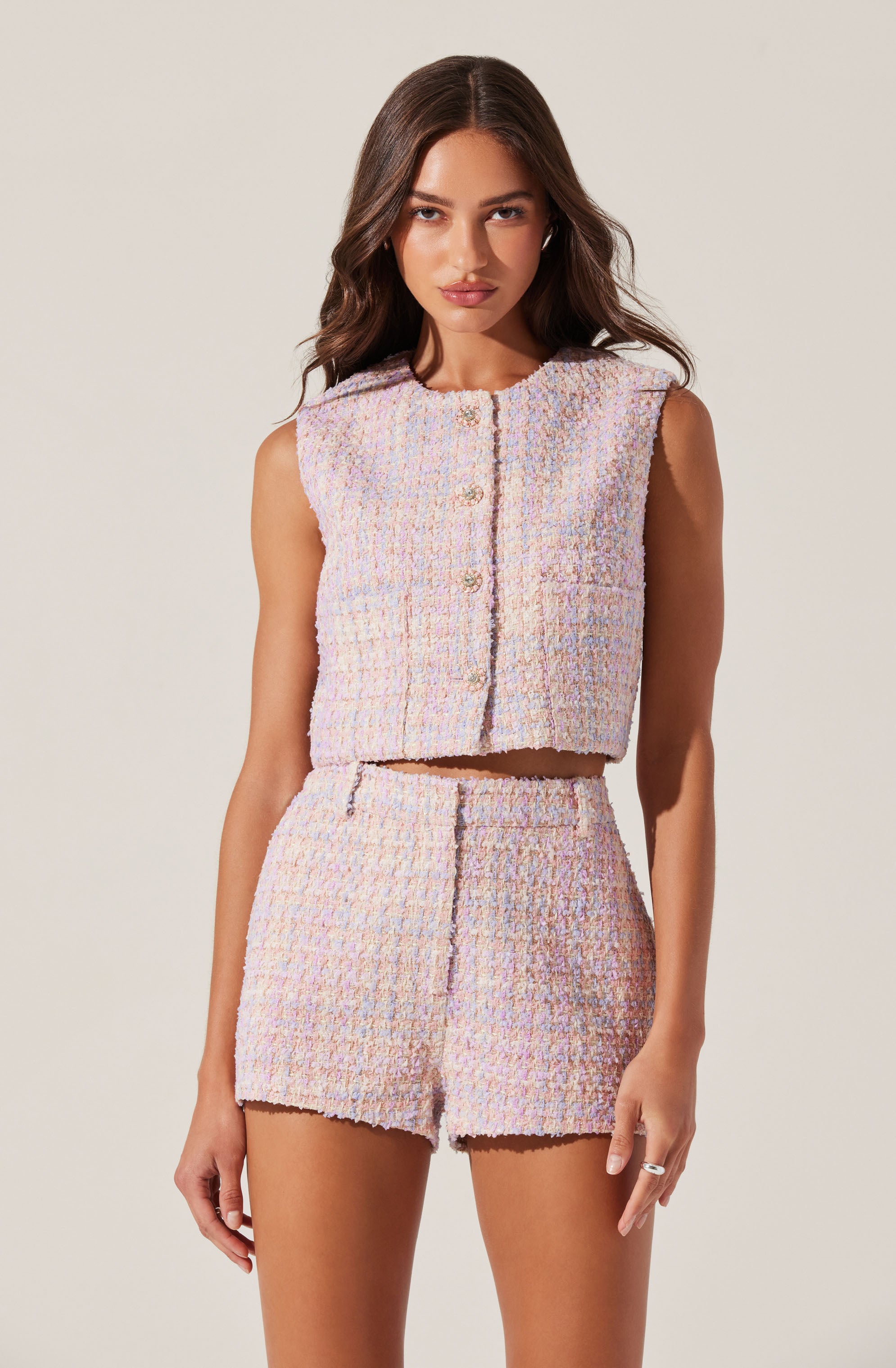 A young woman is wearing a light, pastel-colored, textured sleeveless crop top with a round neckline and button closure down the front. She is also wearing matching high-waisted shorts with the same texture and pastel hues. The outfit has a soft, tweed-like appearance.