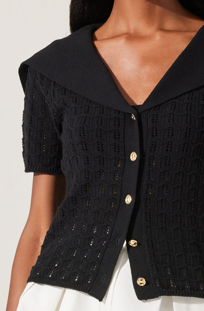 A person is wearing a black, short-sleeved, open-knit cardigan with a textured geometric pattern. The cardigan features a wide collar and gold buttons down the front. They are also wearing white pants.
