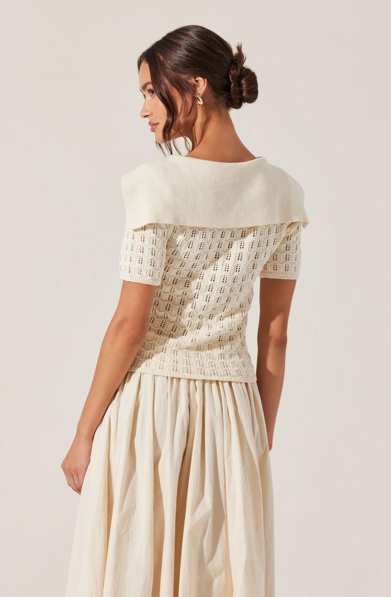 A woman is wearing an off-white, short-sleeved knit top with a wide, boat neckline that drapes over her shoulders. The top features a textured, open-knit pattern. She is also wearing a long, flowing skirt made of a lightweight fabric with a gathered, pleated design in a matching cream color. Her hair is styled in a loose, low bun.