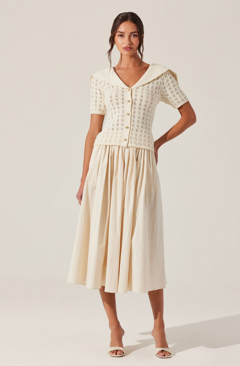 A woman is wearing a cream-colored, short-sleeved, button-up top with a openwork crochet pattern and a wide collar that drapes over her shoulders. She pairs it with a high-waisted, full-length, pleated skirt in a matching cream tone. She is also wearing open-toe heels.