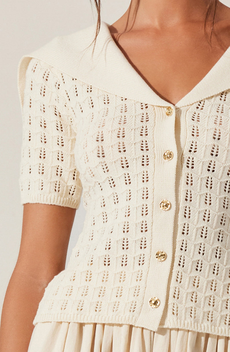 A woman is wearing a cream-colored, short-sleeved, knitted cardigan with a textured open-knit pattern. The cardigan features a wide collar, a button-down front with gold buttons, and is paired with a pleated skirt.