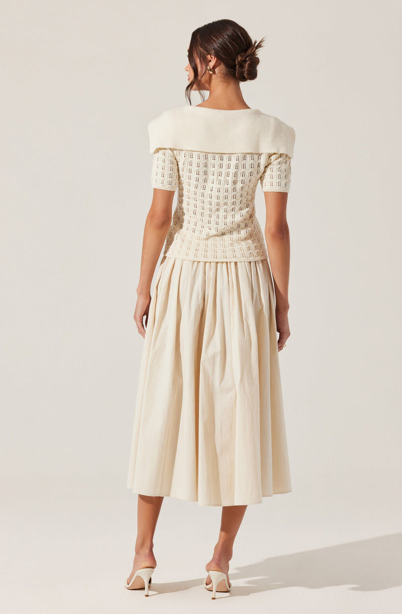 A woman is wearing a white knit top with short sleeves and a wide, draped collar that extends over her shoulders. She pairs this with a long, cream-colored, pleated midi skirt and white high-heeled sandals. Her hair is styled in a low, messy bun.