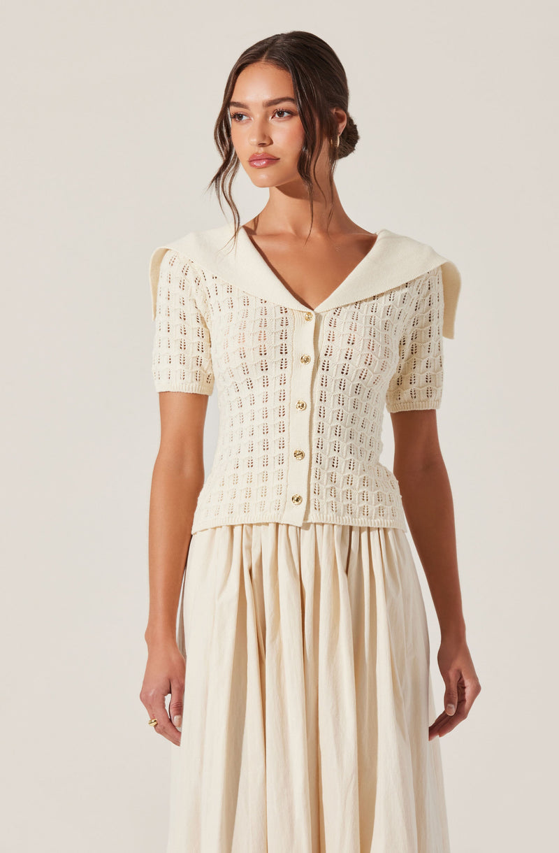 A woman is wearing a cream-colored, short-sleeved, button-up top with a crochet pattern. The top has a wide collar that drapes over the shoulders. She pairs it with a long, flowing cream-colored skirt with gathered fabric, creating a soft, elegant look.
