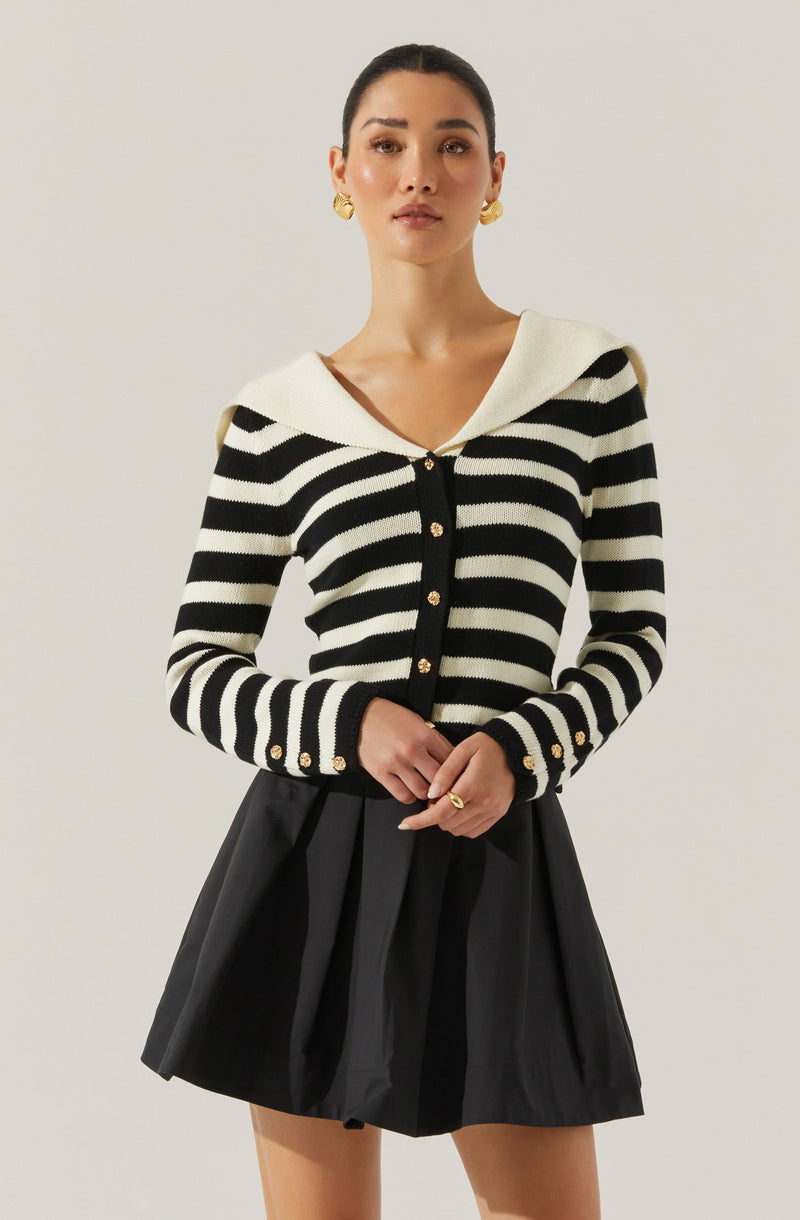 A woman is wearing a long-sleeved, black and white striped cardigan with gold buttons down the front and on the cuffs. The cardigan features a wide, white, sailor-style collar. She pairs it with a high-waisted, black pleated skirt. She has gold hoop earrings and a gold ring on her finger.
