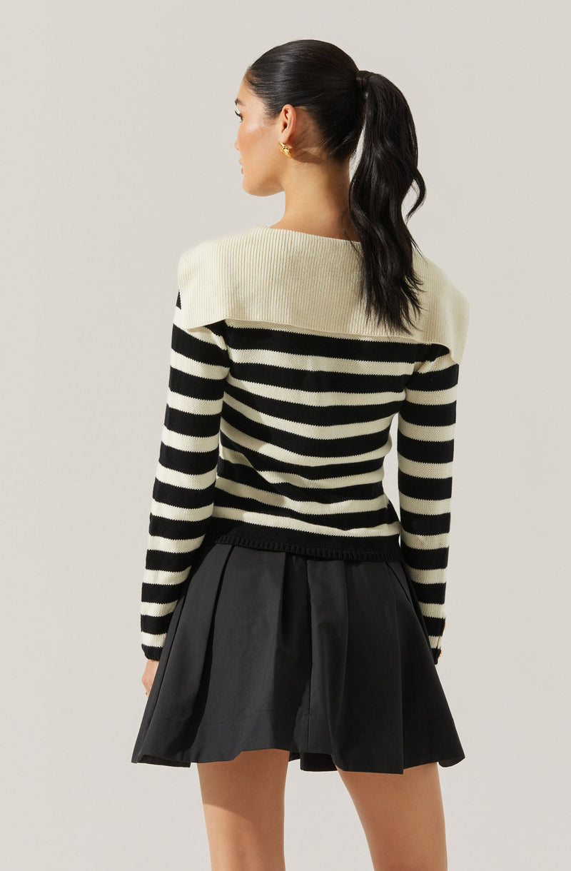 A woman is wearing a cream and black horizontally striped sweater with a wide, structured shoulder detail. The sweater has a ribbed texture and long sleeves. She pairs it with a black, high-waisted, pleated skirt that has a shiny finish.
