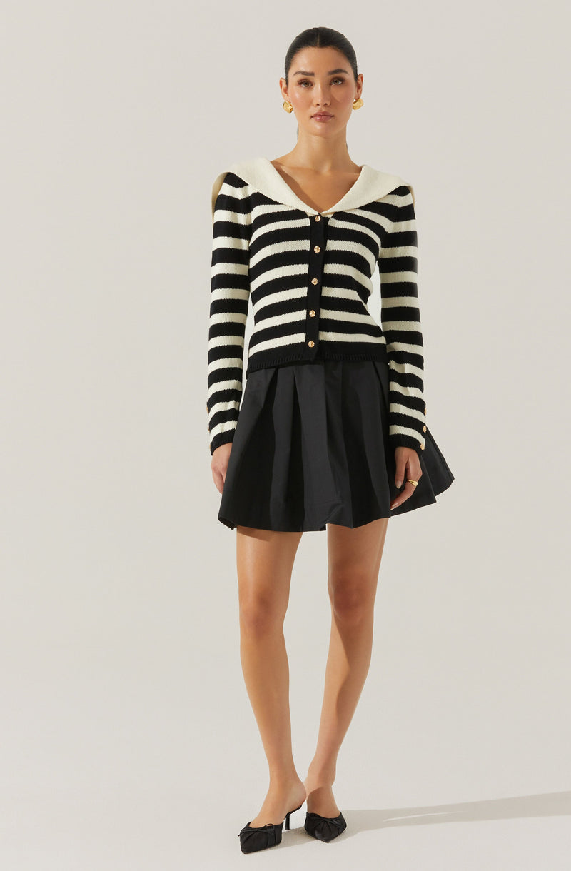 The woman is wearing a black and white horizontal striped long-sleeve cardigan with gold buttons, featuring a wide, white sailor-style collar. She pairs this with a black pleated skirt and black pointed-toe shoes with bows on the front. She is also holding a black clutch in her left hand and accessorized with gold earrings and rings.