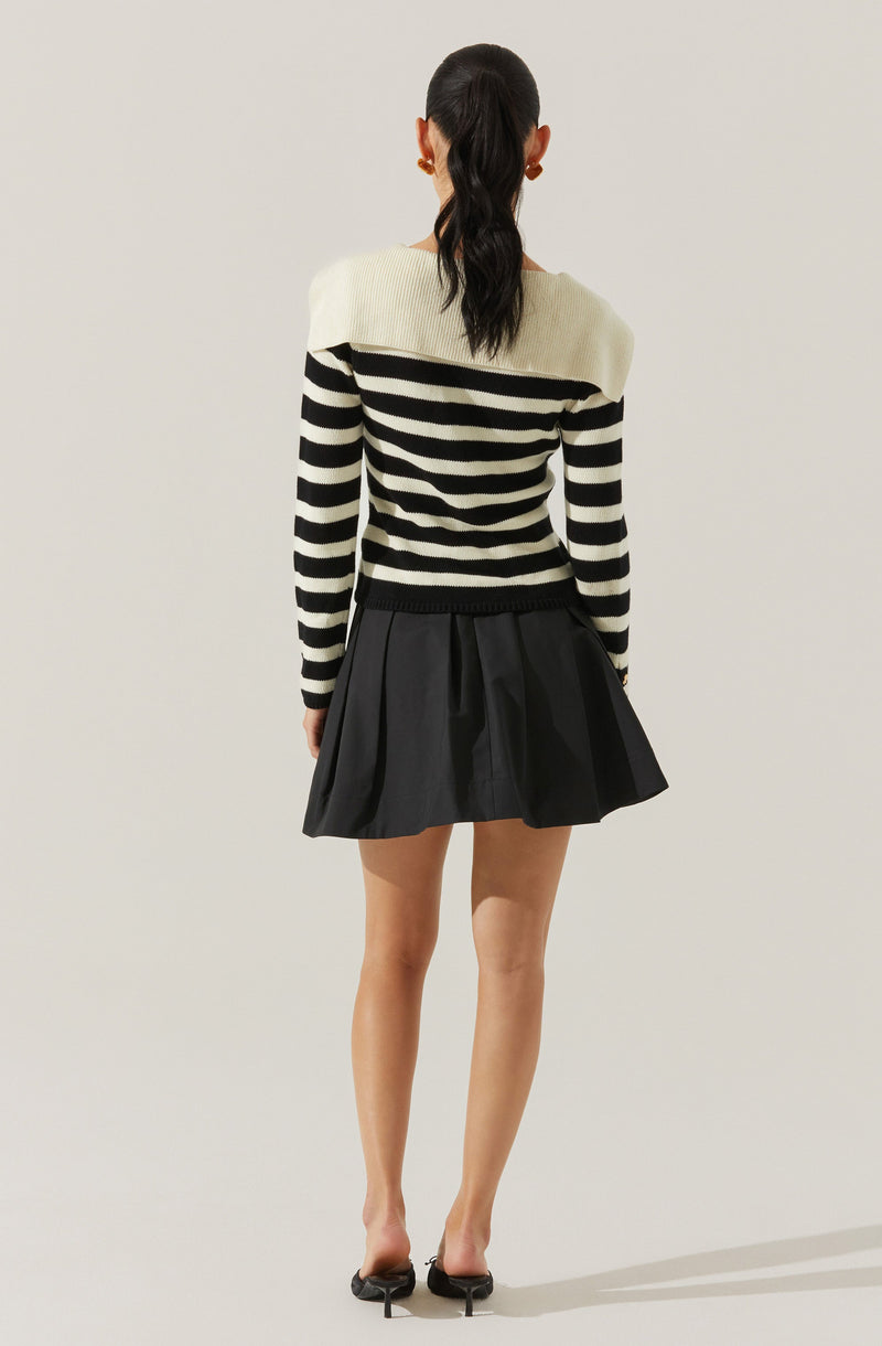 A person is wearing a long-sleeve sweater with black and white horizontal stripes. The sweater has white shoulders and black stripes on the chest, arms, and lower part. They are also wearing a high-waisted, pleated, black skirt that flares out above the knees. The person has black high-heeled shoes.