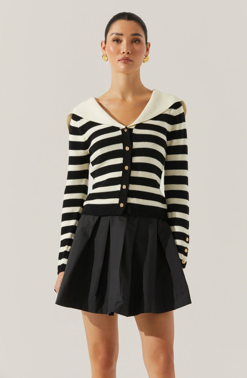 A woman is wearing a long-sleeve, black and white striped cardigan with a wide, white, folded collar. The cardigan features a row of gold buttons down the front and on the cuffs. She is also wearing a black skirt with pleats.