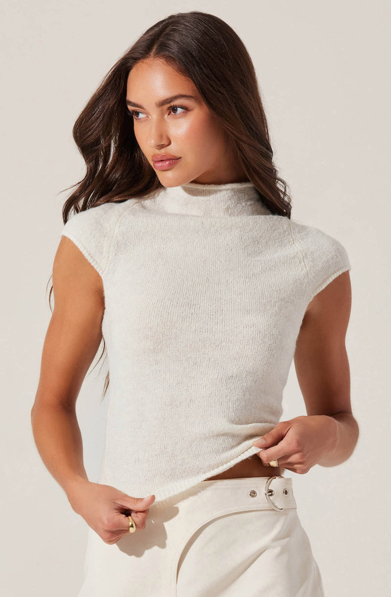 A woman is wearing a sleeveless, off-white knit top with a high, rounded neckline. She pairs it with white high-waisted pants that have a belt. The outfit is simple and elegant, with a neutral color palette.