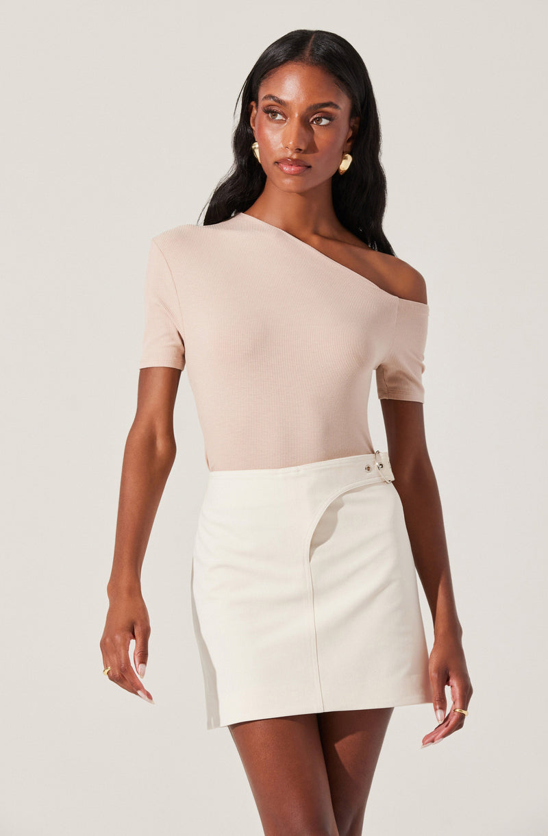 A woman is wearing a light pink, off-the-shoulder top with short sleeves and a fitted silhouette. She pairs it with a high-waisted, cream-colored mini skirt that features a decorative belt with metal accents on the side. She accessorizes with gold earrings and rings.
