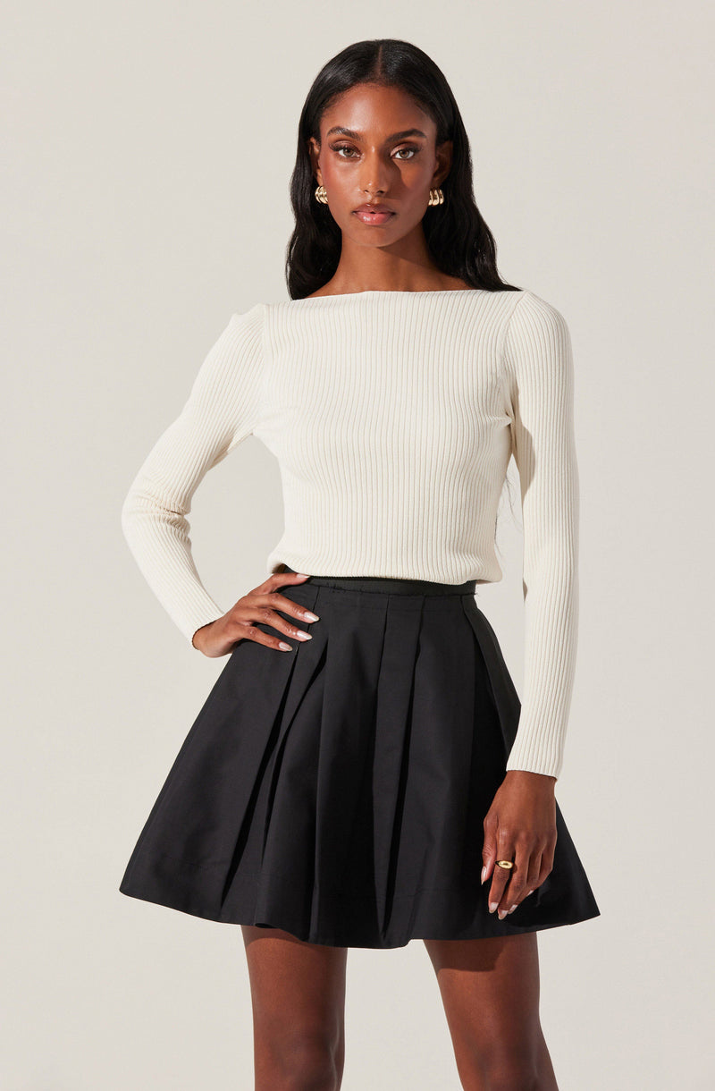 A woman is wearing a long-sleeved, cream-colored, ribbed sweater with a boat neckline, paired with a high-waisted, black A-line skirt featuring pleats.