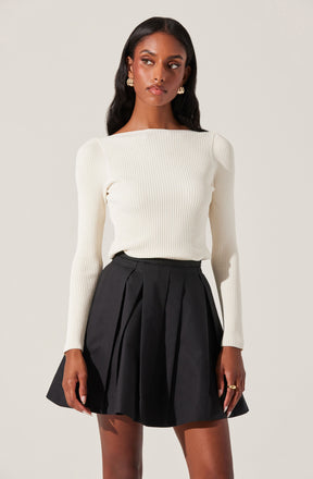 A woman is wearing a fitted, long-sleeved, ribbed sweater in an off-white or cream color. She pairs it with a high-waisted black skirt that has pleats and a flared silhouette. She accessorizes with gold earrings and rings.