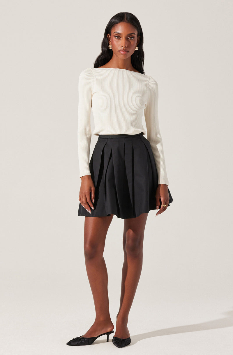 A woman is wearing a long-sleeved, cream-colored ribbed sweater with a boat neckline paired with a high-waisted black skirt that has pleats. She is accessorized with gold hoop earrings, and her feet are in black pointed-toe flats with small bows at the front.