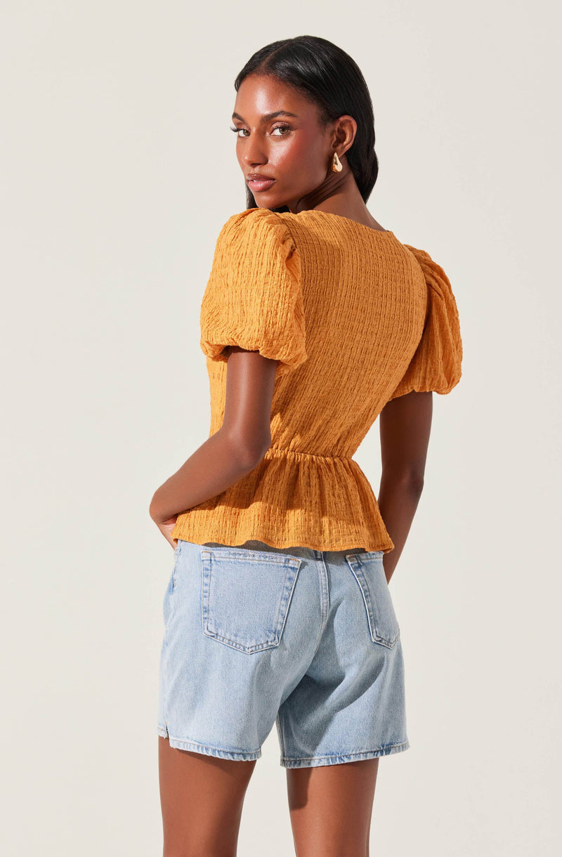 A woman is wearing a mustard yellow, textured, short-sleeve blouse with a peplum hem and puffed sleeves. She is also wearing light-wash denim shorts with a relaxed fit.