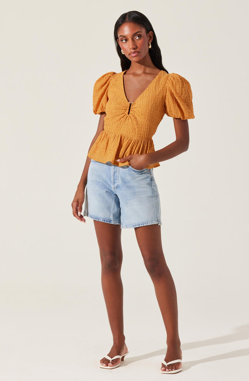 A woman is wearing a mustard yellow top with puffed short sleeves and a fitted, textured bodice featuring a front zipper and a peplum hem. She pairs it with light blue denim shorts and white heeled sandals.