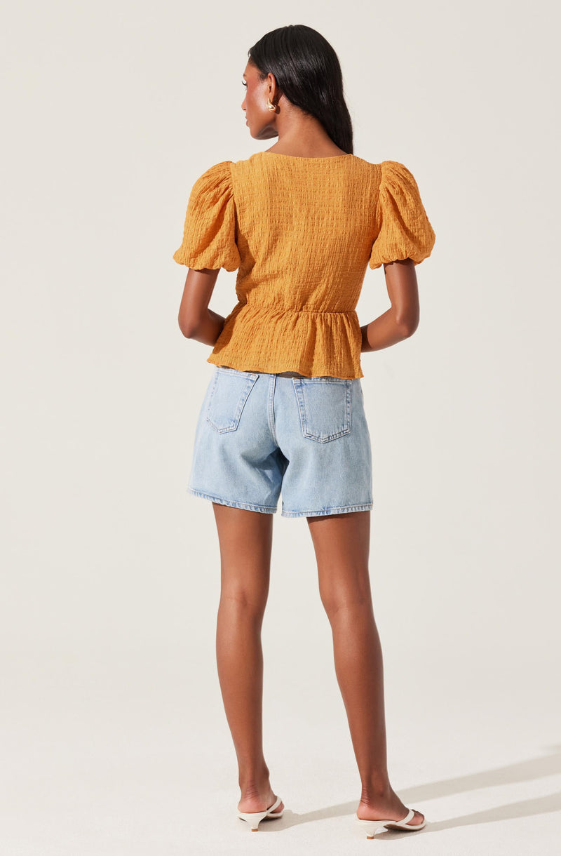 The woman is wearing a mustard yellow top with puffed short sleeves and a gathered waist, made of a textured fabric. She pairs it with light wash denim shorts that have a relaxed fit and two back pockets. She is also wearing a pair of white open-toe flats.