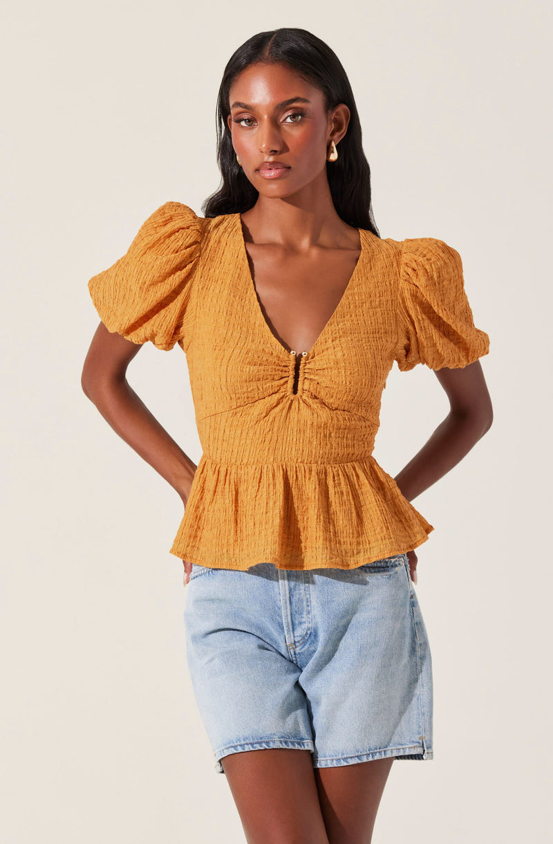 A woman is wearing a mustard yellow top with puffed short sleeves, a V-neckline with a small zipper detail in the center, and a peplum hem. The fabric of the top appears textured and slightly crinkled. She is also wearing light denim shorts.