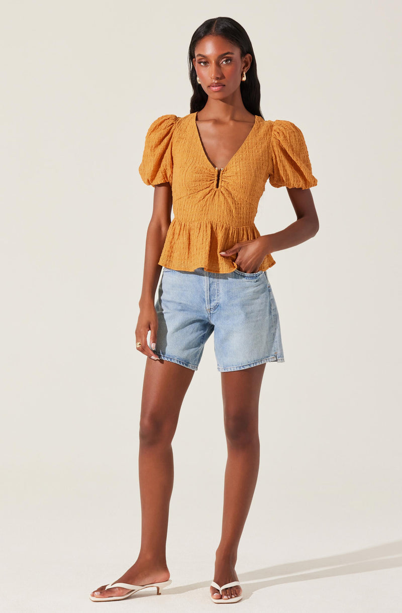 A woman is wearing a mustard yellow blouse with a textured fabric, short puff sleeves, and a V-neckline with a front zipper. The blouse has a gathered waistline with a ruffled hem. She pairs it with light blue, slightly distressed denim shorts and beige flat sandals.