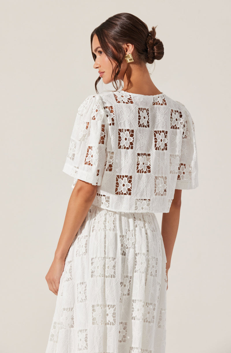 A woman is wearing a matching white lace outfit with a short-sleeved, cropped top and a high-waisted, floor-length skirt. The lace fabric features intricate square and floral embroidery patterns. She accessorizes with gold geometric earrings and has her dark hair styled in a low, twisted bun.