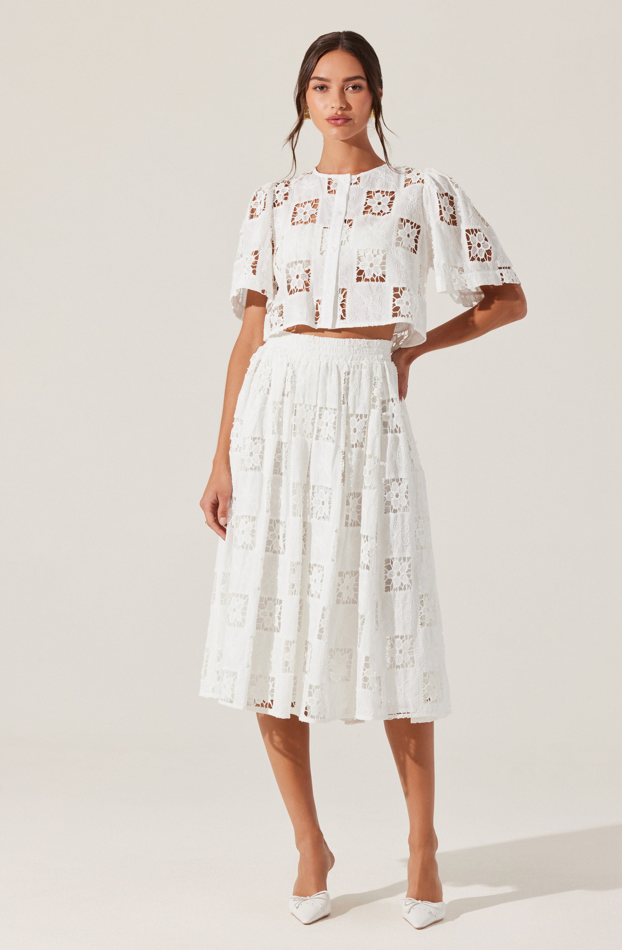 A woman is wearing a white cropped top and a matching white midi skirt, both made from fabric with a geometric cut-out pattern. The top has short sleeves and buttons down the front, while the skirt is high-waisted with a relaxed fit. She is also wearing white pointed-toe shoes.