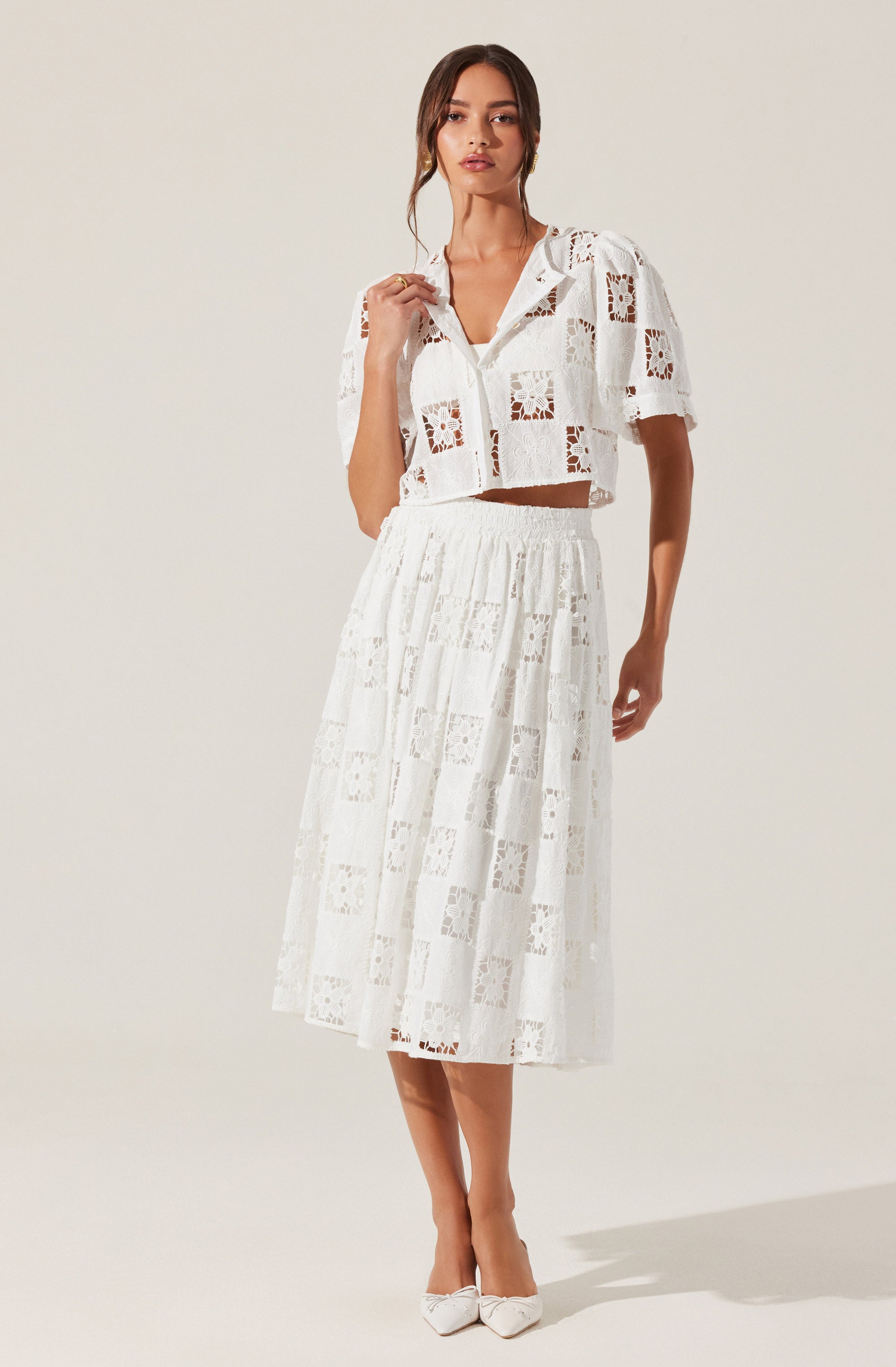 A woman is wearing a white, two-piece outfit made of lace fabric. The top is a short-sleeved, collared shirt with a button-down front and a cropped length, featuring a pattern of square and floral lace designs. The skirt is a high-waisted, midi-length skirt with a similar lace pattern, reaching below the knees. She is also wearing white shoes with a pointed toe.