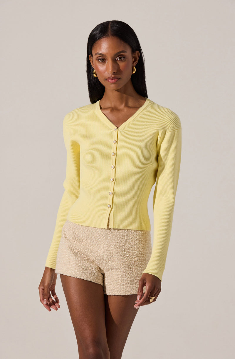 A woman is wearing a light yellow, long-sleeve, V-neck cardigan with matching buttons down the front. She pairs it with beige textured shorts. She has dark, straight hair and is accessorized with gold hoop earrings and a gold ring.