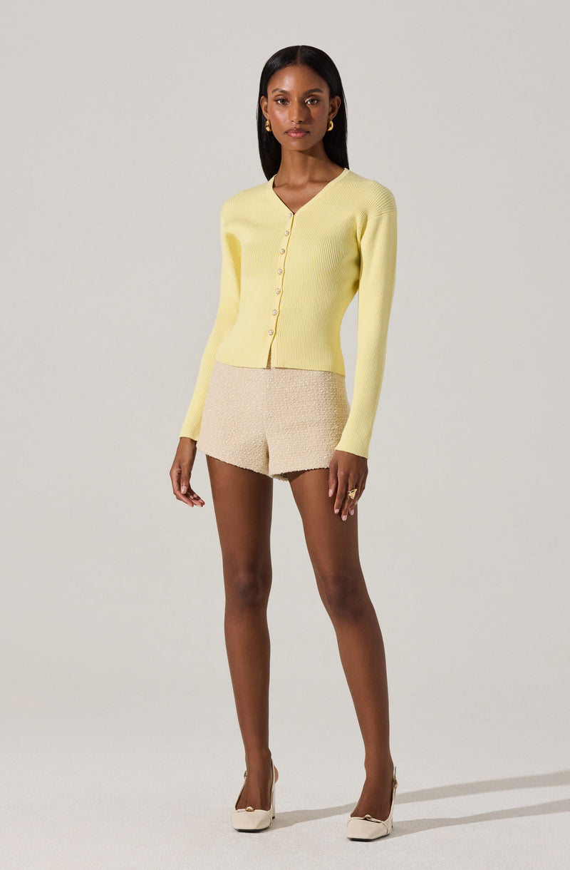 A woman is wearing a light yellow, long-sleeved, V-neck cardigan with buttons down the front. She pairs it with beige textured shorts and beige high-heeled shoes. She has minimal jewelry, including gold hoop earrings.