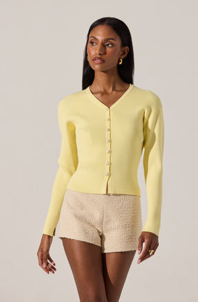 A woman is wearing a light yellow, ribbed, long-sleeve cardigan with a V-neckline and pearl-like buttons down the front. She has paired it with beige textured shorts.