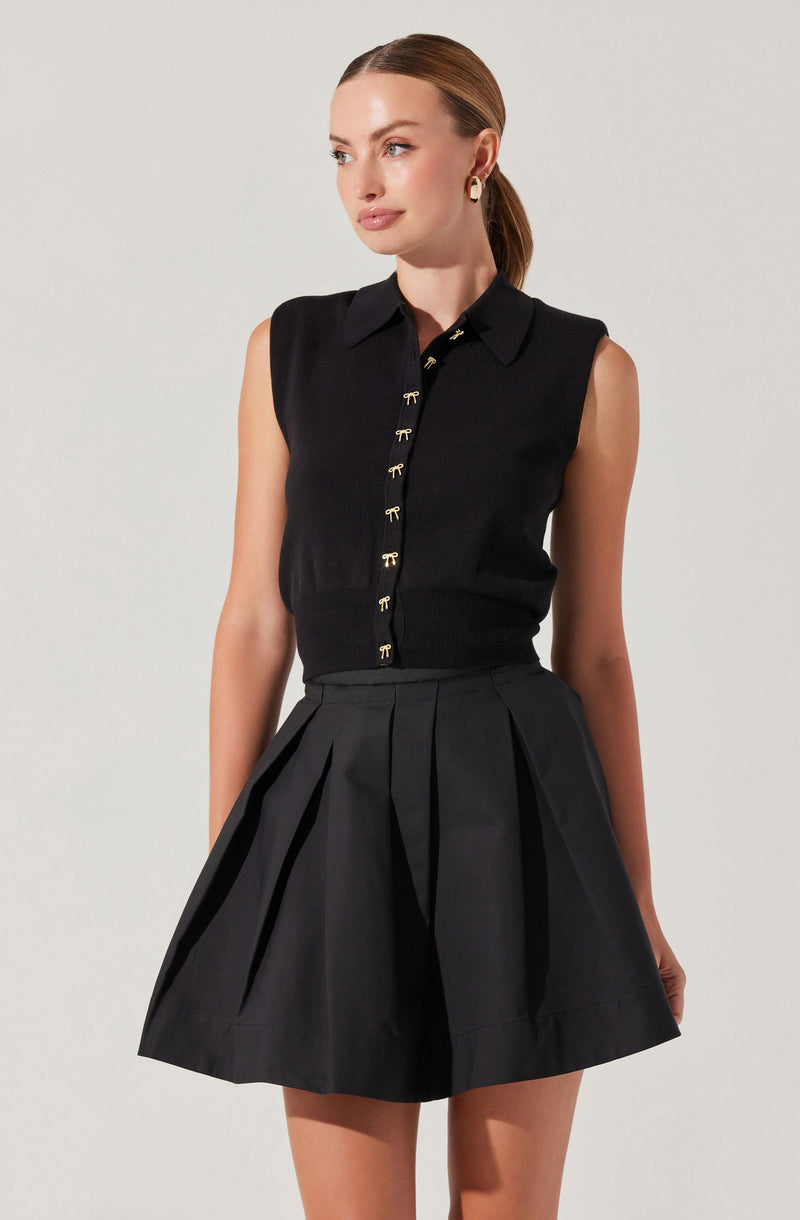 A woman is wearing a sleeveless black collared shirt with gold bow-shaped buttons down the front, paired with a high-waisted black pleated skirt.