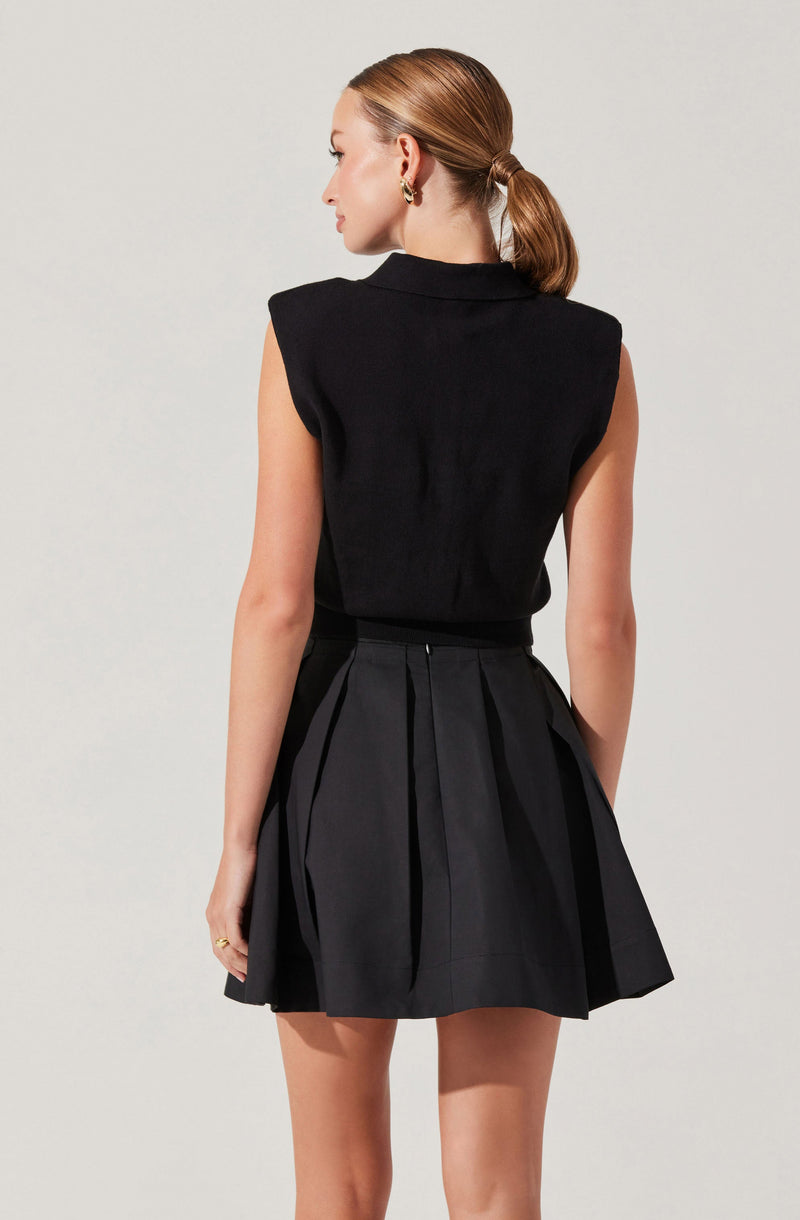 A woman with her hair tied back in a low ponytail is wearing a sleeveless black top with padded shoulders and a high collar. She pairs this with a high-waisted black pleated skirt. She is also wearing gold hoop earrings and a ring on her left hand.