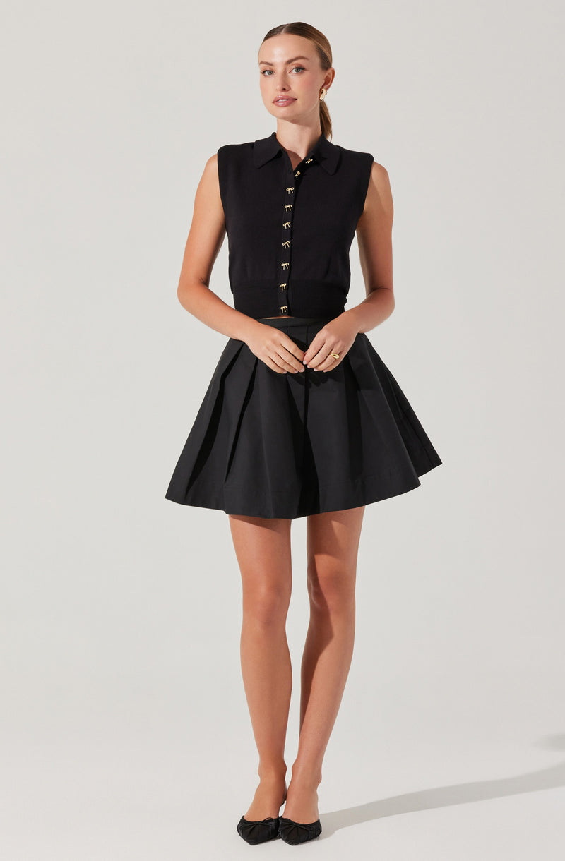 A woman is wearing a sleeveless black top with a collar and gold-tone button details down the front. She pairs it with a high-waisted, black, pleated knee-length skirt. She completes the outfit with black flats that have a bow on the front.