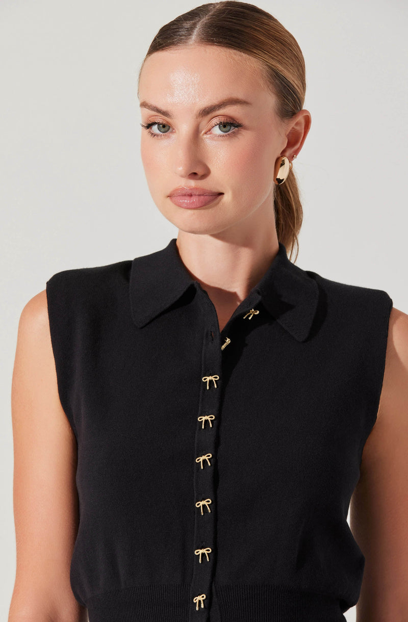 A woman is wearing a sleeveless black knit top with a collared neckline. The top features small gold bow-shaped embellishments along the front placket. She has gold hoop earrings and her hair is styled in a sleek, low ponytail.