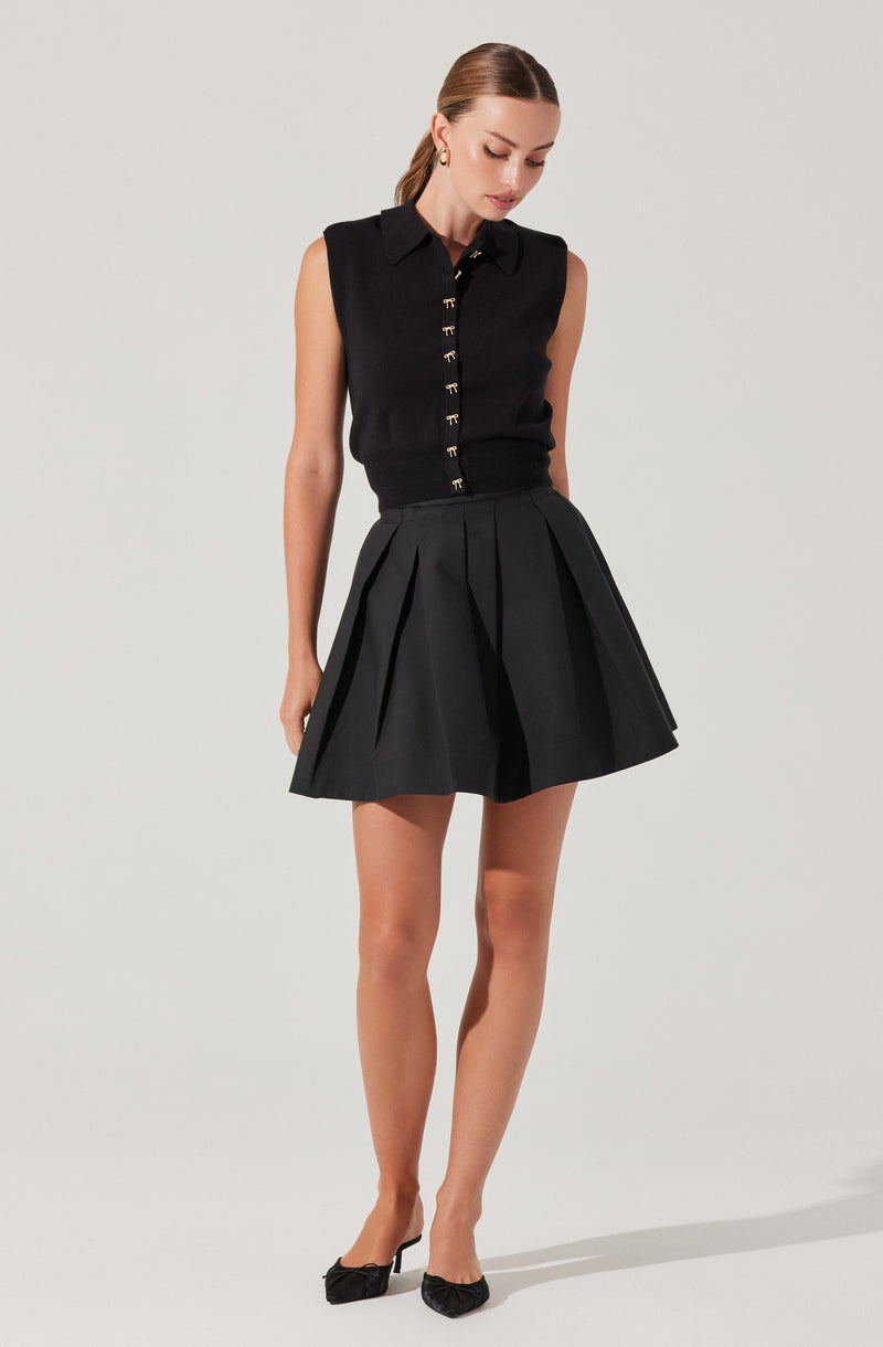 A woman is wearing a sleeveless black blouse with a collar and gold button details down the front. She pairs it with a high-waisted black pleated skirt that falls above the knees. She completes her outfit with black heels and small gold hoop earrings.