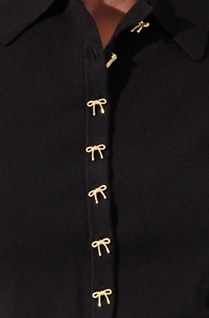 A close-up of a black garment with a row of small, gold bow-shaped buttons down the front. The fabric appears to be smooth and slightly textured.