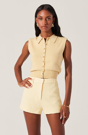 A woman is wearing a sleeveless, cream-colored button-up top with a collar. She pairs it with matching high-waisted shorts that have a textured fabric. She accessorizes with gold earrings, a bracelet, and a ring.