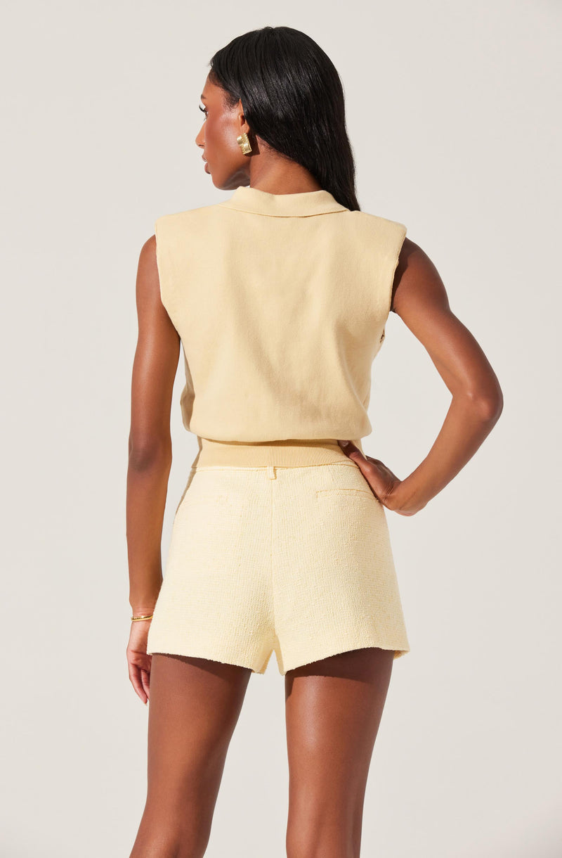 A woman is wearing a sleeveless, light yellow top with a structured shoulder design and a collared neckline, paired with matching high-waisted shorts made of a textured fabric.