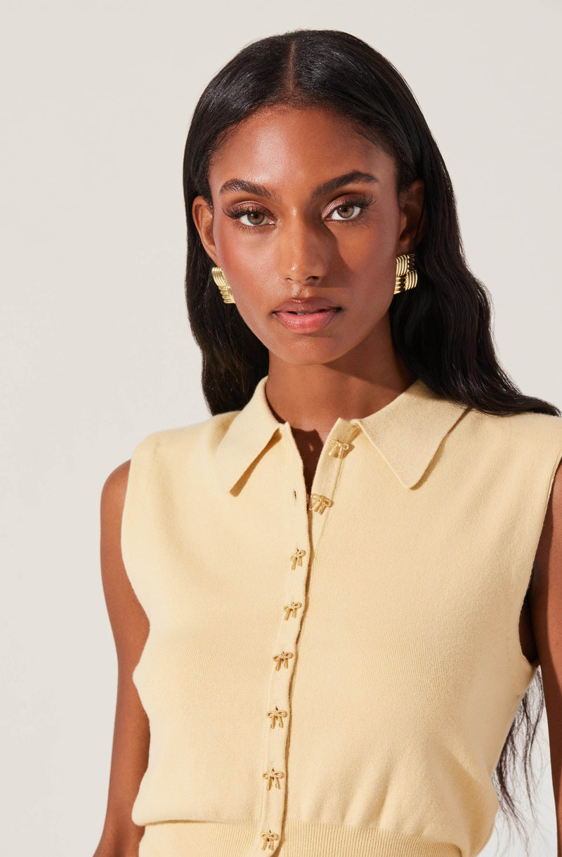 A woman is wearing a sleeveless, pale yellow cardigan with a collar and small bow-shaped buttons down the front. She has large, gold, hoop earrings with a textured pattern.