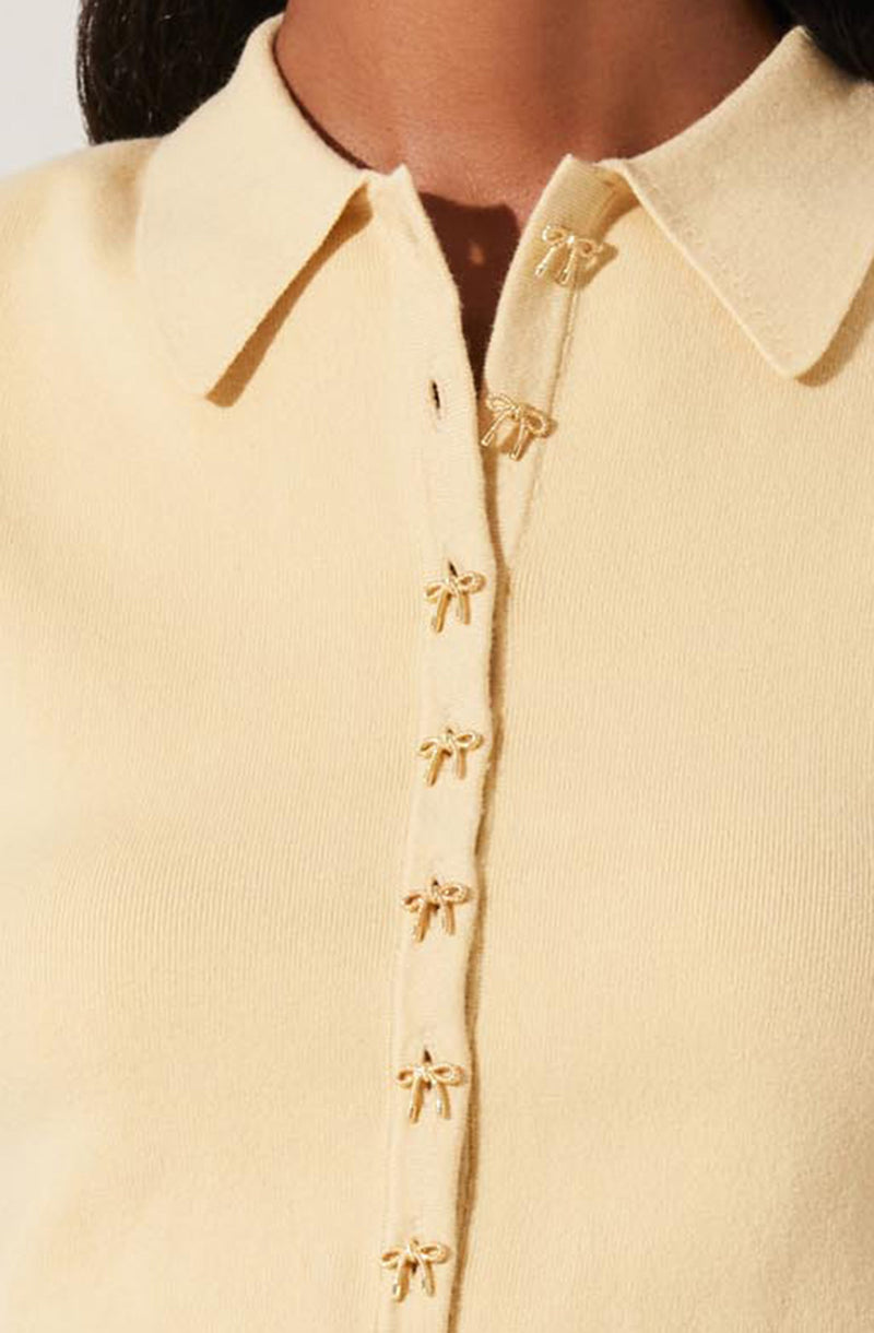 A close-up of a light yellow collared shirt featuring a row of small decorative bows along the front placket. The shirt has a classic shirt collar and a zipper closure underneath the bows.
