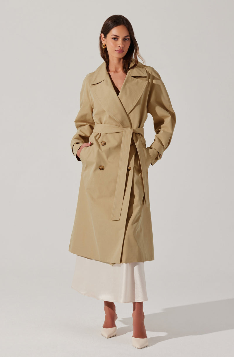 A woman is wearing a classic beige trench coat with a belted waist, double-breasted buttons, and a wide lapel. The coat has long sleeves and side pockets. She pairs it with a white skirt and white high-heeled shoes. She also has small gold earrings.