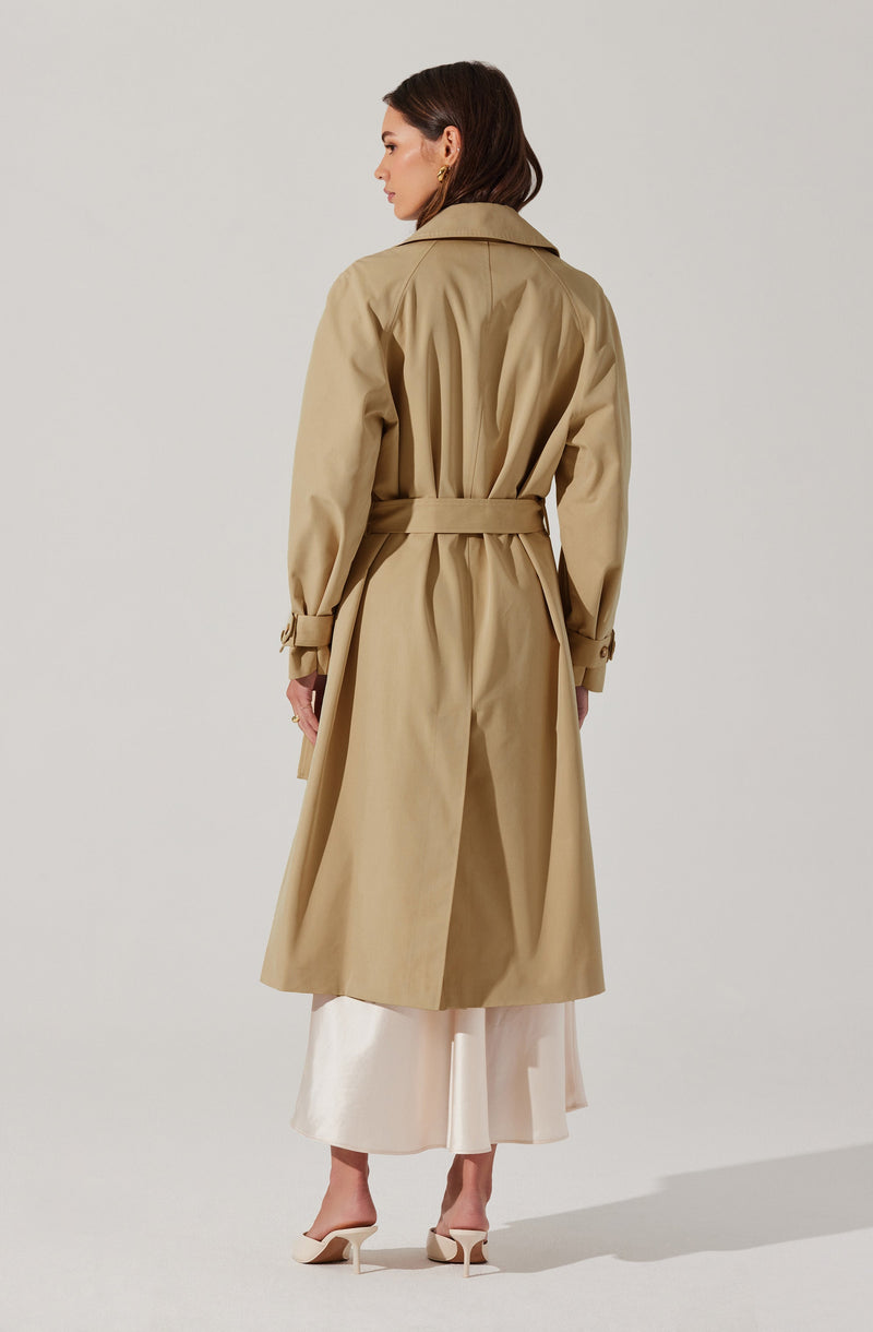 A woman is wearing a long beige trench coat with a belt at the waist, buttoned cuffs on the sleeves, and a classic collar. She has paired the coat with a light-colored, ankle-length dress and beige high-heeled shoes. The coat's design includes pleats and a vent at the back.