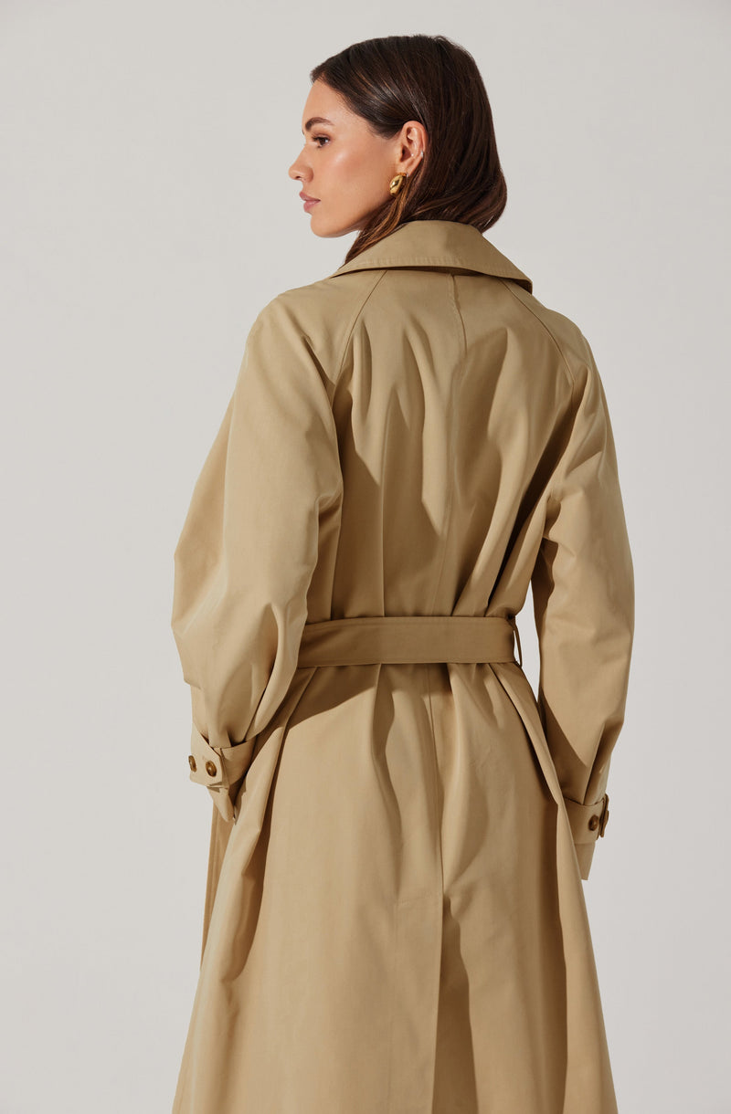 A woman is wearing a beige trench coat with a wide collar, long sleeves with buttoned straps at the cuffs, and a matching belt cinched at the waist. The coat features a classic design with a smooth, slightly shiny fabric.