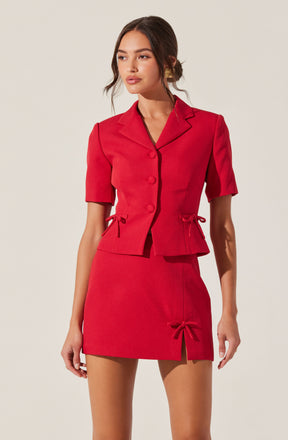 A woman is wearing a coordinated red outfit consisting of a tailored short-sleeve blazer with a notched collar and front button closure, and a matching high-waisted mini skirt with a front slit and decorative bow ties on the sides and front. The blazer features a fitted silhouette with darts at the waist, and the skirt is form-fitting. She has long, dark hair styled in loose waves and is accessorized with a pair of gold earrings.