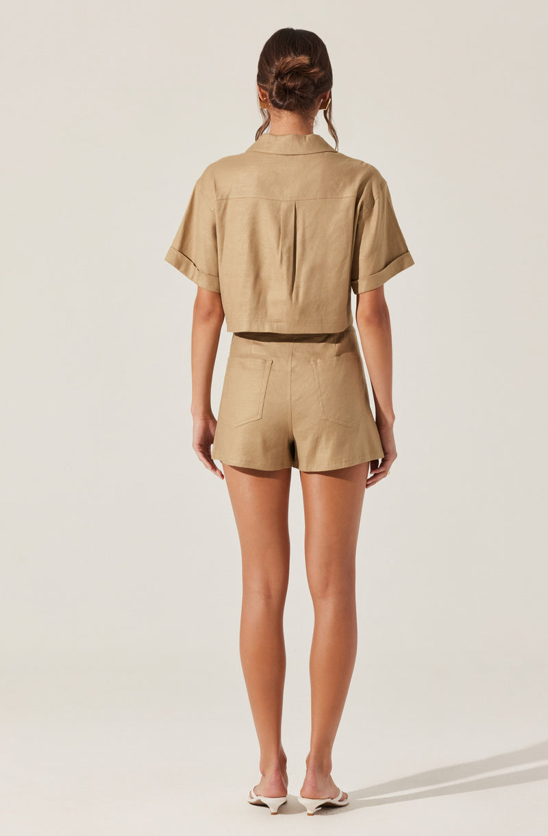 A woman is wearing a beige short-sleeved button-up shirt with a collar and a back yoke, paired with matching high-waisted shorts that have a tailored fit and a single back pocket. She has her hair styled in a loose bun and is standing against a plain, light-colored background.