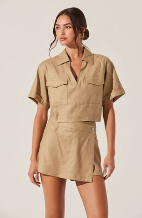 A woman is wearing a beige short-sleeved shirt with a collar, featuring two chest pockets with flap closures. She pairs it with a matching beige wrap skirt that has a side slit and a button detail.