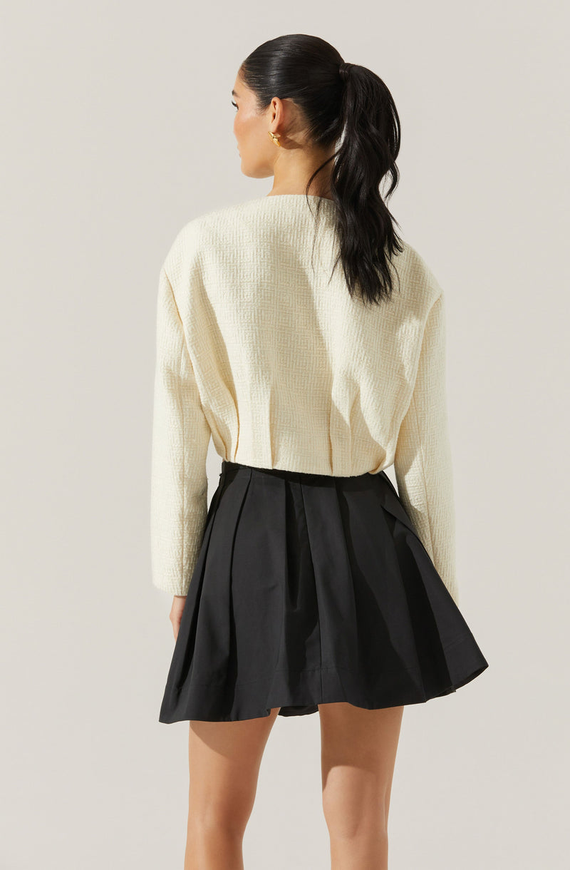 A woman is wearing an oversized, cream-colored knit sweater with a textured pattern, paired with a high-waisted, black pleated skirt that has a slightly flared silhouette. Her dark hair is styled in a high ponytail.