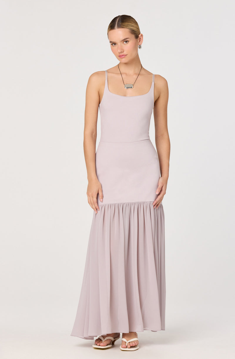 A woman is wearing a sleeveless, light-colored dress with thin straps. The top part of the dress fits closely to her body, while the skirt features a gathered, flowing section that extends to the floor. She is also wearing a necklace with a rectangular pendant and simple earrings.